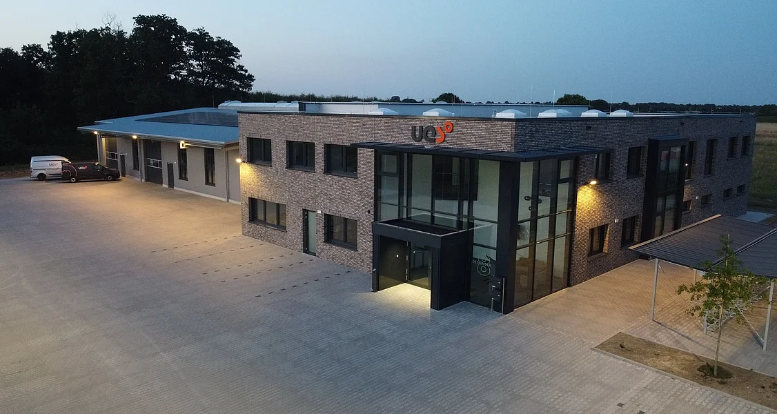 Luftaufnahme eines modernen zweigeschossigen Bürogebäudes mit beleuchtetem Eingangsbereich und Firmenlogo an der Fassade bei Dämmerung