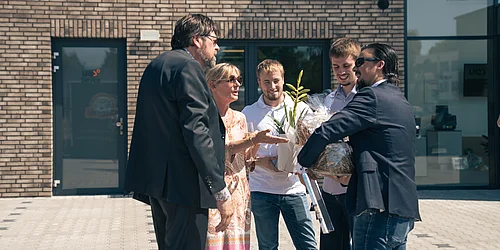 Freudige Begrüßung! V.l.n.r.: Carsten Weiner, Assistenz und rechte Hand, Sonja Weiler, sowie Lars Pätow, Jannis Wrampelmeier, John Frank von JB. Fünf Personen im Freien vor einem modernen Gebäude, zwei Männer übergeben einem Paar und einer Frau einen großen, eingepackten Geschenkkorb und eine Pflanze