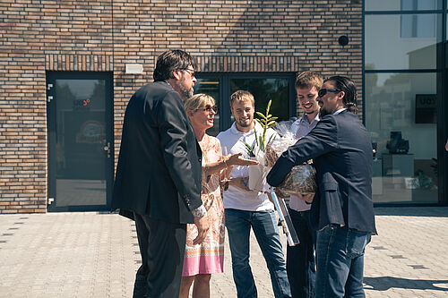 Freudige Begrüßung! V.l.n.r.: Carsten Weiner, Assistenz und rechte Hand, Sonja Weiler, sowie Lars Pätow, Jannis Wrampelmeier, John Frank von JB. Fünf Personen im Freien vor einem modernen Gebäude, zwei Männer übergeben einem Paar und einer Frau einen großen, eingepackten Geschenkkorb und eine Pflanze