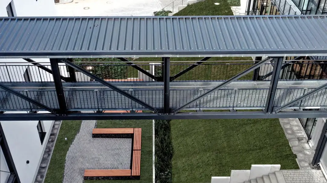 Blick von oben auf eine überdachte Fußgängerbrücke zwischen zwei Gebäuden, darunter Treppen, gepflasterte Wege, Rasenflächen und mehrere geparkte Autos.