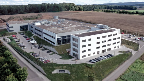 Luftaufnahme eines modernen, weißen Bürogebäudes mit mehreren Parkplätzen, umgeben von Feldern und Wald.