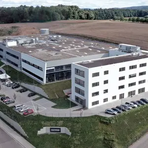 Luftaufnahme eines modernen, weißen Bürogebäudes mit mehreren Parkplätzen, umgeben von Feldern und Wald.