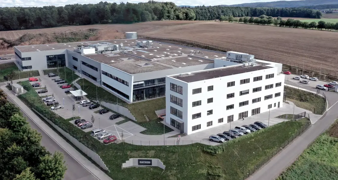 Luftaufnahme eines modernen, weißen Bürogebäudes mit mehreren Parkplätzen, umgeben von Feldern und Wald.