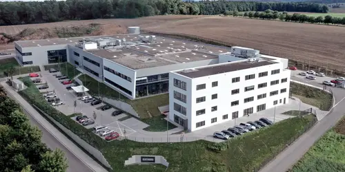 Luftaufnahme eines modernen, weißen Bürogebäudes mit mehreren Parkplätzen, umgeben von Feldern und Wald.