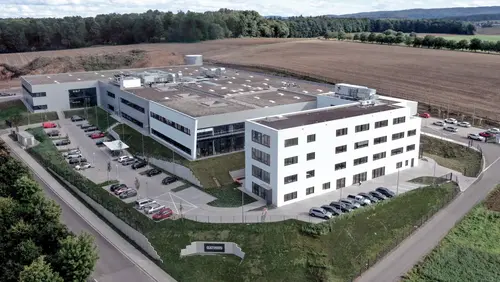 Luftaufnahme eines modernen, weißen Bürogebäudes mit mehreren Parkplätzen, umgeben von Feldern und Wald.