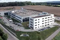 Luftaufnahme eines modernen, weißen Bürogebäudes mit mehreren Parkplätzen, umgeben von Feldern und Wald.