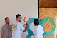 Drei Männer stehen vor einer großen blauen Skulptur aus ineinandergreifenden Ringen, einer hält einen Hammer und schlägt auf einen Holzklotz, im Hintergrund eine Leinwand und eine Wand aus Holzplatten