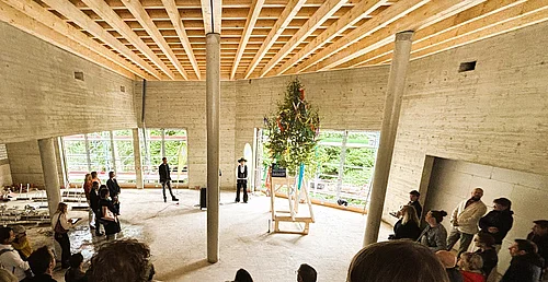 Menschen stehen im Kreis in einem großen Raum mit sichtbarer Holzbalkendecke um einen geschmückten Richtbaum auf einem Podest