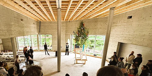 Menschen stehen im Kreis in einem großen Raum mit sichtbarer Holzbalkendecke um einen geschmückten Richtbaum auf einem Podest
