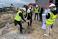 Mehrere Personen mit Schutzhelmen und Warnwesten stehen um eine geöffnete Grube auf einer Baustelle.