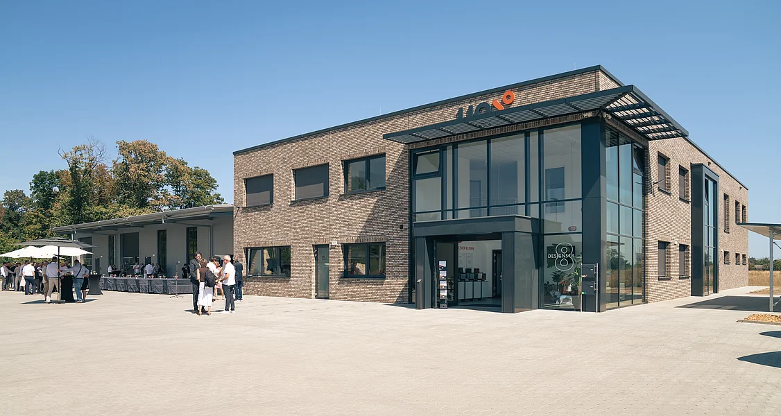 Modernes zweigeschossiges Gebäude mit Glasfassade und Backsteinmauerwerk vor sonnigem Himmel und Menschen auf gepflastertem Vorplatz