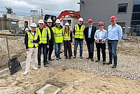 Gruppe von Menschen mit Bauhelmen und Warnwesten steht auf einer Baustelle vor einem Gebäude und einem Bagger