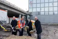 Vier Personen in Winterkleidung führen einen symbolischen Spatenstich auf einer Baustelle vor einem modernen Gebäude durch