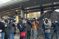 Gruppe von Pressevertretern mit Kameras und Mikrofonen vor einer Baustelle mit Erdhaufen und Betonbrücke