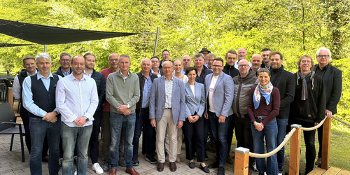 Gruppe von Personen in legerer Kleidung steht auf gepflastertem Platz vor grünem Waldhintergrund