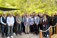 Gruppe von Personen in legerer Kleidung steht auf gepflastertem Platz vor grünem Waldhintergrund