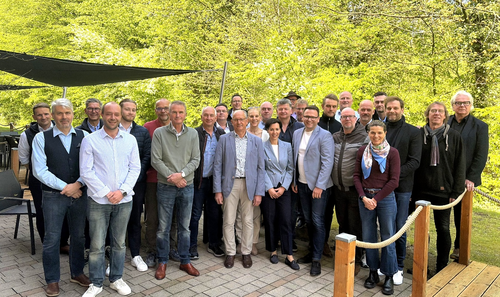 Gruppe von Personen in legerer Kleidung steht auf gepflastertem Platz vor grünem Waldhintergrund