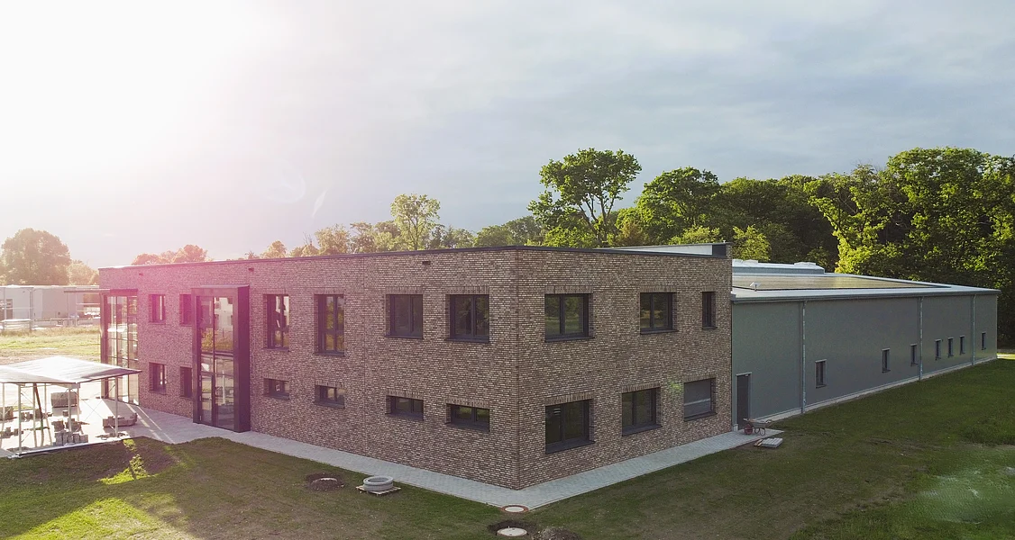 Neubau einer Produktionshalle mit Büro Flaches, zweigeschossiges Gebäude aus hellen Ziegeln mit mehreren Fenstern und angrenzendem langgestrecktem Anbau auf einer grünen Wiese bei Sonnenlicht