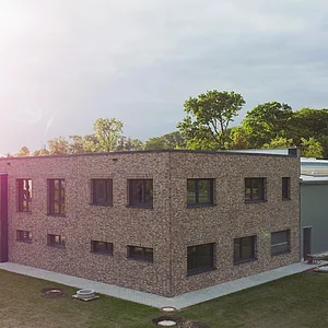Neubau einer Produktionshalle mit Büro Flaches, zweigeschossiges Gebäude aus hellen Ziegeln mit mehreren Fenstern und angrenzendem langgestrecktem Anbau auf einer grünen Wiese bei Sonnenlicht