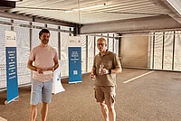 Dankbare Idee bei 39°C: Marc Johannes brachte Eis und Eiswürfel für uns und die Besucher vorbei! Zwei Männer stehen in einem Parkhaus, einer hält ein Getränk, der andere ein Papier.