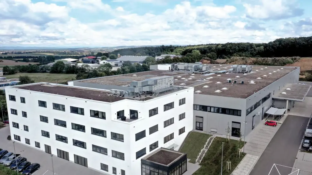 Luftaufnahme eines weißen, mehrstöckigen Verwaltungsgebäudes neben einer großen Halle mit Parkplätzen und umliegender Landschaft.