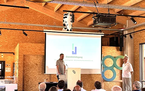 Mann steht am Rednerpult vor Leinwand mit Präsentation zum Thema Grundsteinlegung, Publikum sitzt im Vordergrund, zweiter Mann steht rechts neben einer großen Skulptur mit zwei ineinander verschlungenen Ringen