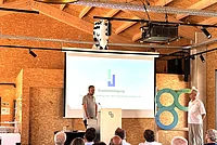Mann steht am Rednerpult vor Leinwand mit Präsentation zum Thema Grundsteinlegung, Publikum sitzt im Vordergrund, zweiter Mann steht rechts neben einer großen Skulptur mit zwei ineinander verschlungenen Ringen