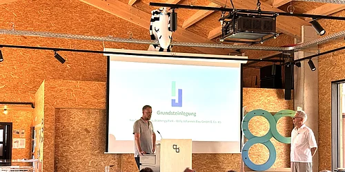 Mann steht am Rednerpult vor Leinwand mit Präsentation zum Thema Grundsteinlegung, Publikum sitzt im Vordergrund, zweiter Mann steht rechts neben einer großen Skulptur mit zwei ineinander verschlungenen Ringen