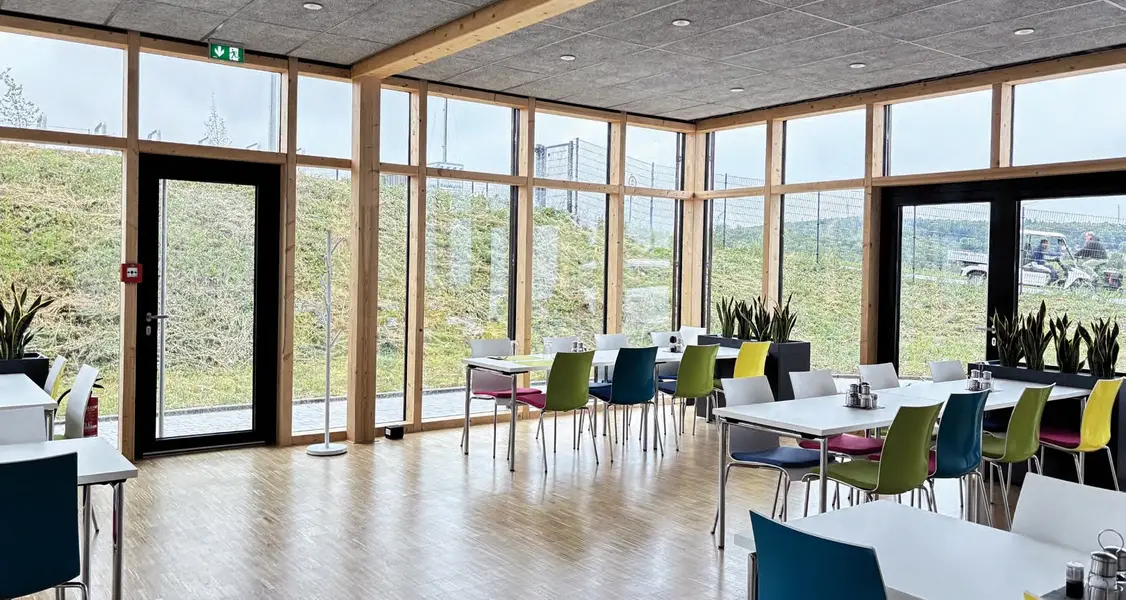 Großer Wintergarten mit bodentiefen Fenstern, mehreren weißen Tischen und bunten Stühlen in Blau, Grün und Gelb.