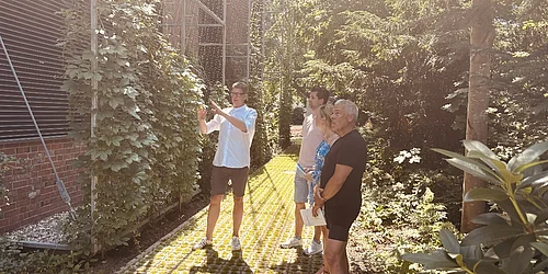 Ein Bild wie aus den Tropen: Lars Gevers und Falk Hildebrandt erläutern die aufwendige Grünfassade. Vier Personen stehen im Garten, eine zeigt mit ausgestrecktem Arm auf eine mit Kletterpflanzen bewachsene Wand