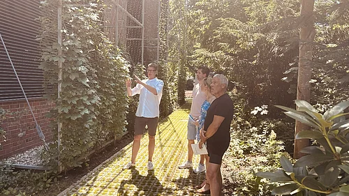 Vier Personen stehen im Garten, eine zeigt mit ausgestrecktem Arm auf eine mit Kletterpflanzen bewachsene Wand