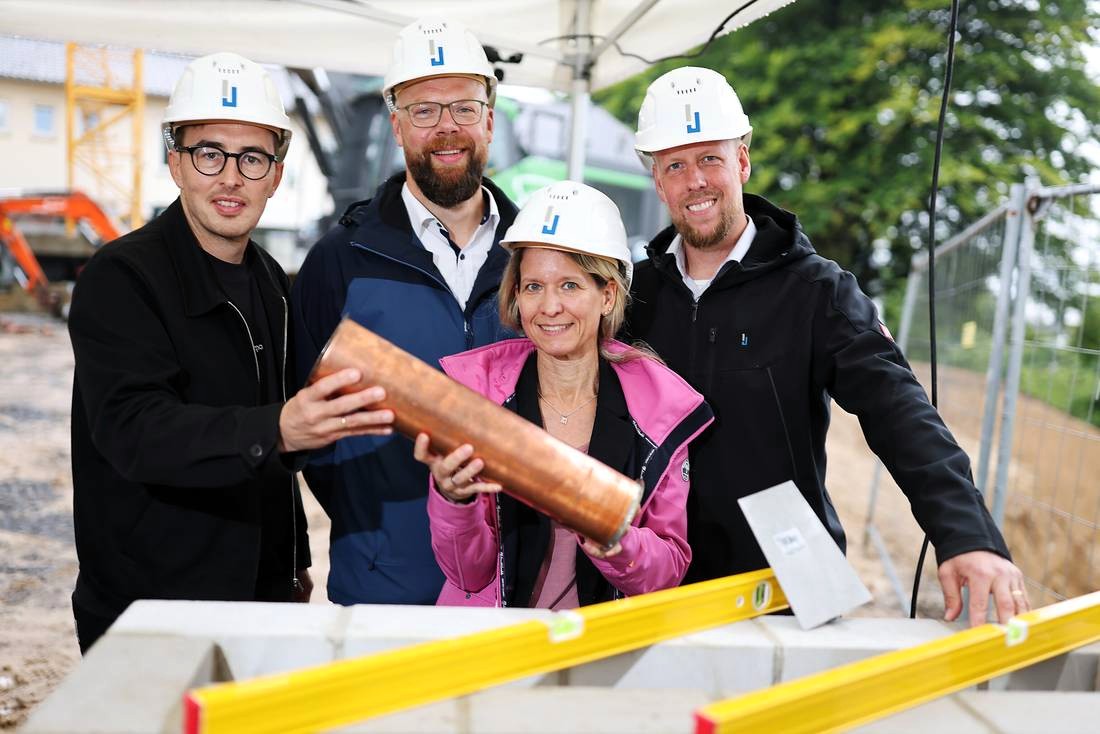 Vier Personen mit Bauhelmen halten gemeinsam eine Metallröhre über einer Mauer auf einer Baustelle